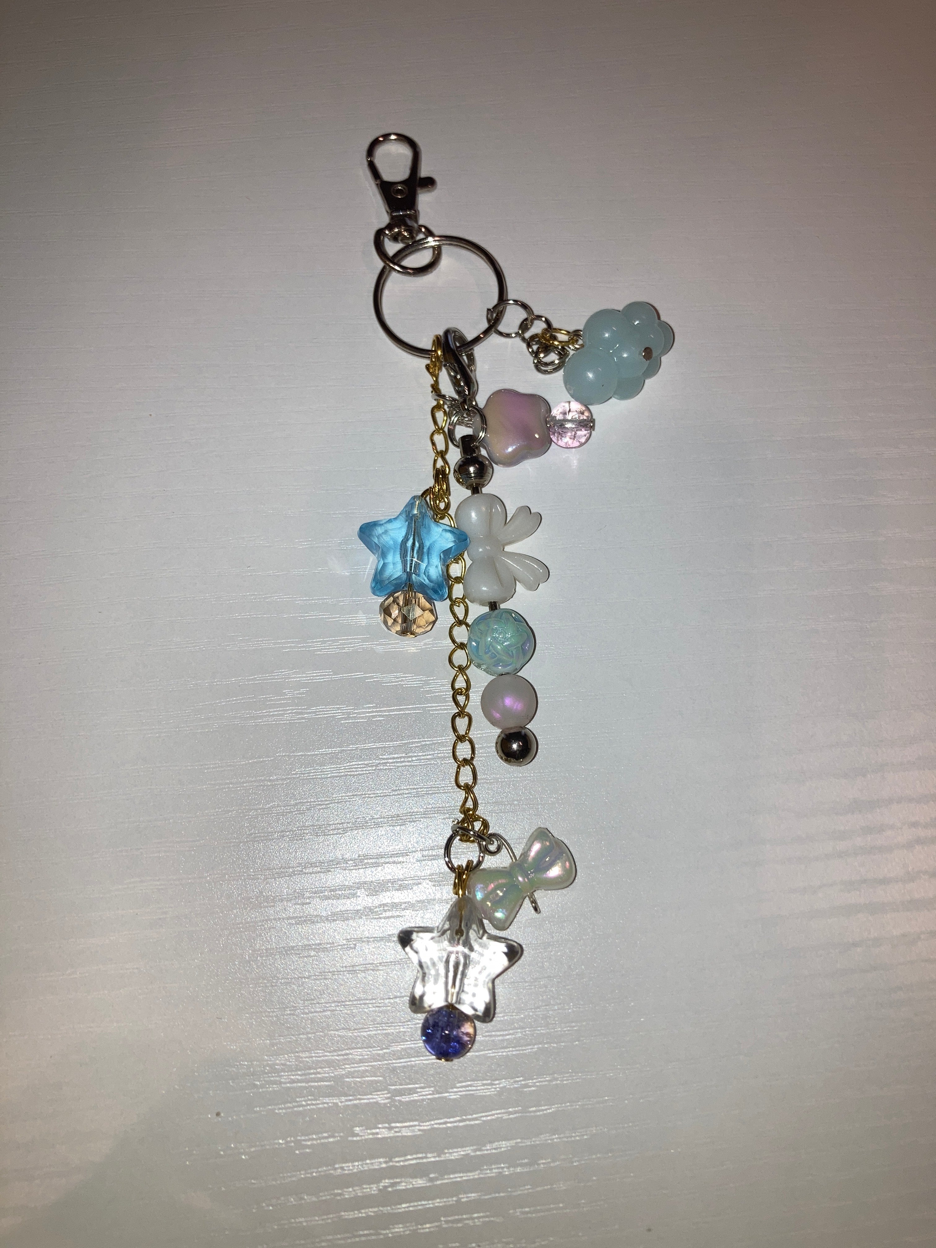 A gold-tone chain keychain charm featuring pastel-colored beads, translucent flower charms, a blue star, and a purple crystal, attached to a silver lobster clasp on a white background.