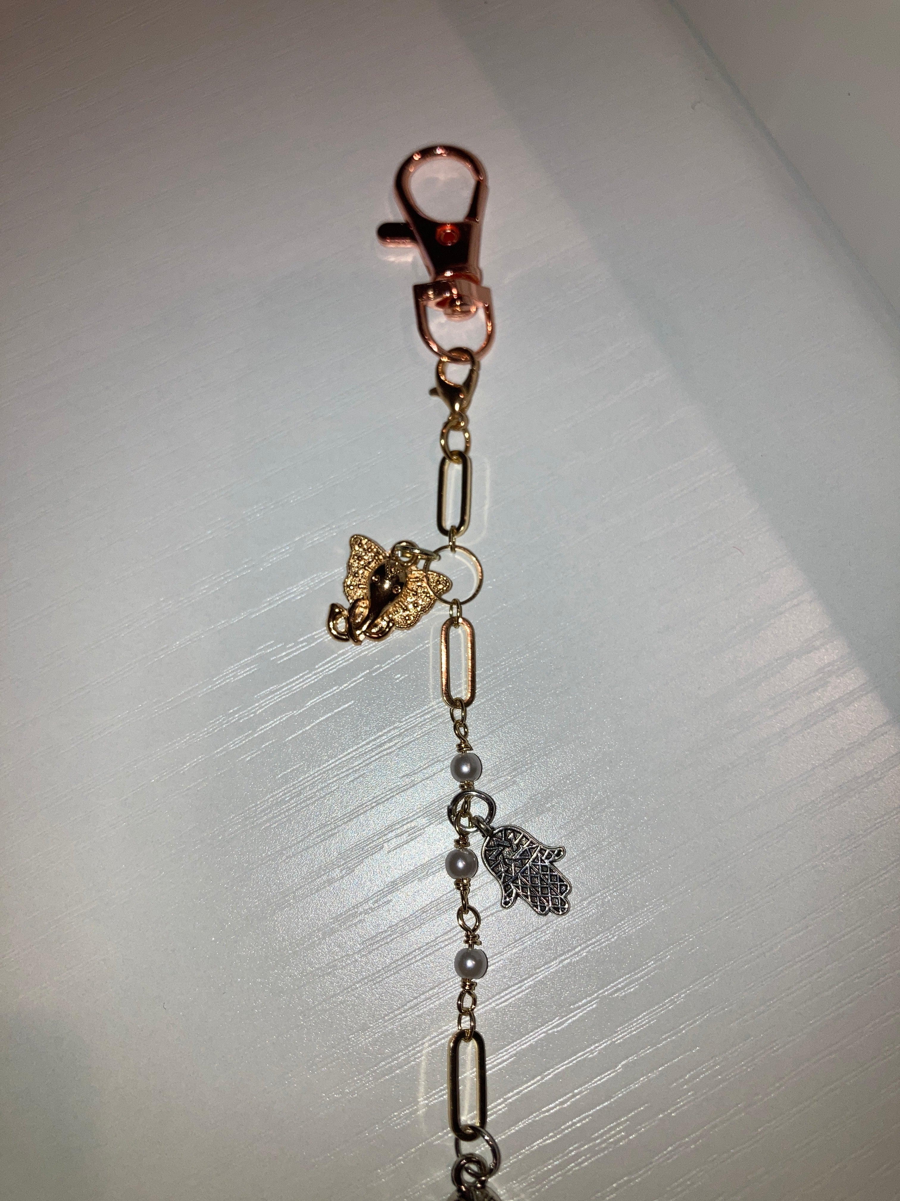 A close-up of a jewelry accessory featuring a rose gold lobster clasp, a gold elephant charm, silver and rose gold paperclip links, gray pearl beads, and a silver hamsa hand charm, displayed on a light gray textured surface.