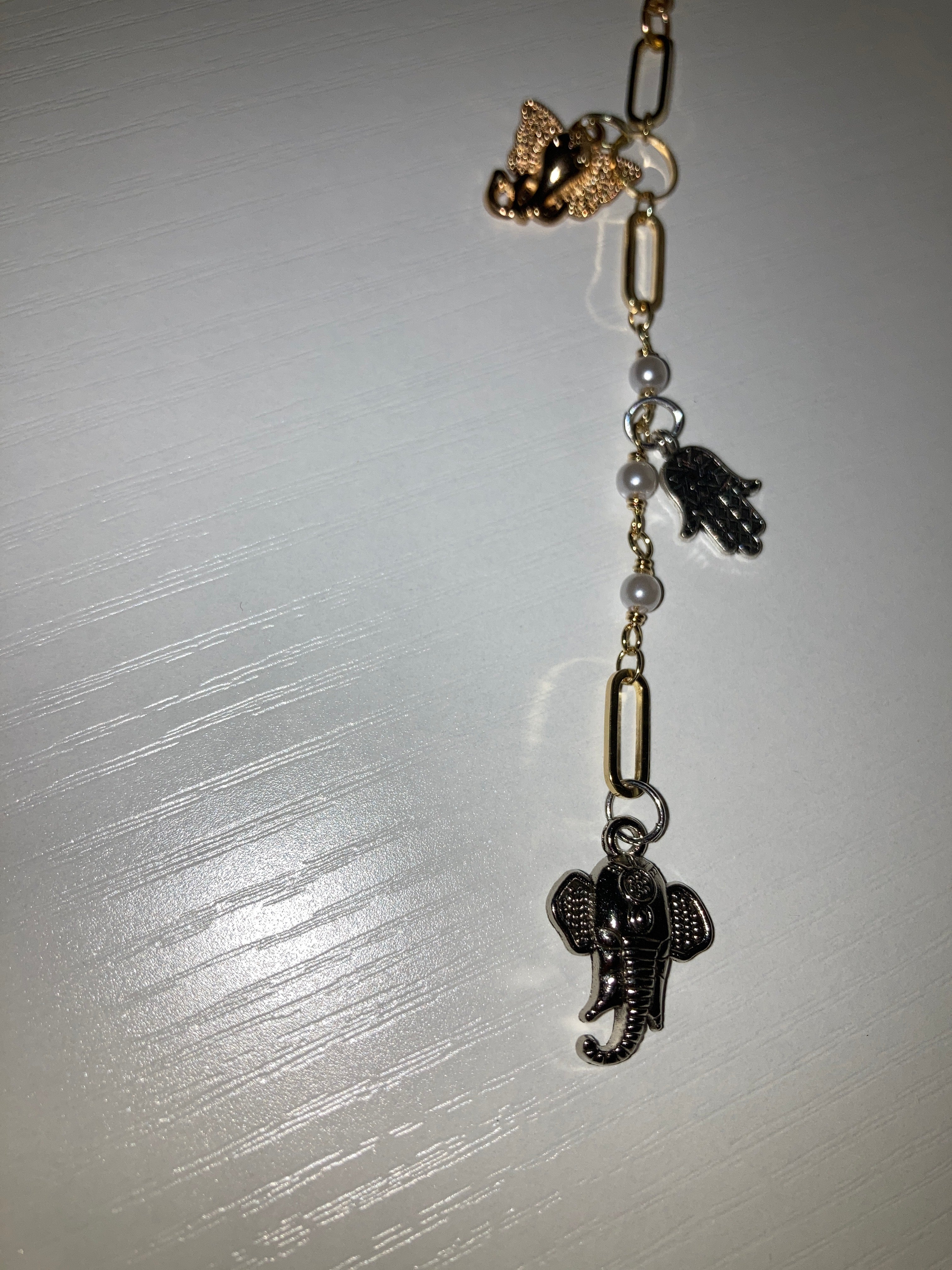 A close-up of a jewelry accessory featuring a rose gold lobster clasp, a gold elephant charm, silver and rose gold paperclip links, gray pearl beads, and a silver hamsa hand charm, displayed on a light gray textured surface.