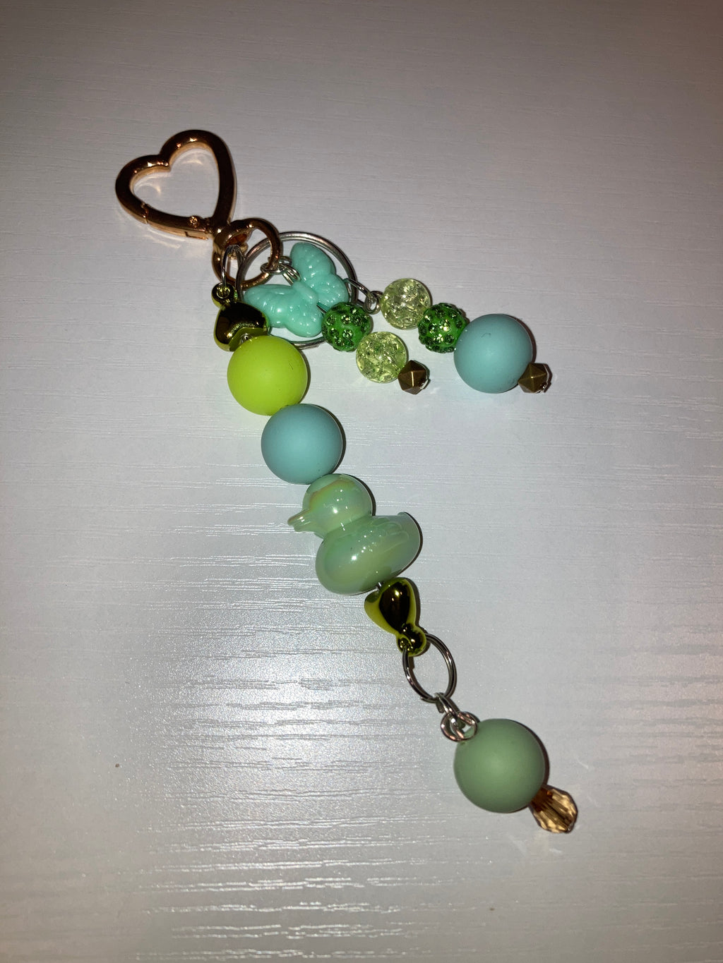 A beaded keychain or bag charm with pastel light blue, mint green, and lime green beads, clear faceted beads, a small gold heart charm, and a gold-tone metal ring, shown on a white background.