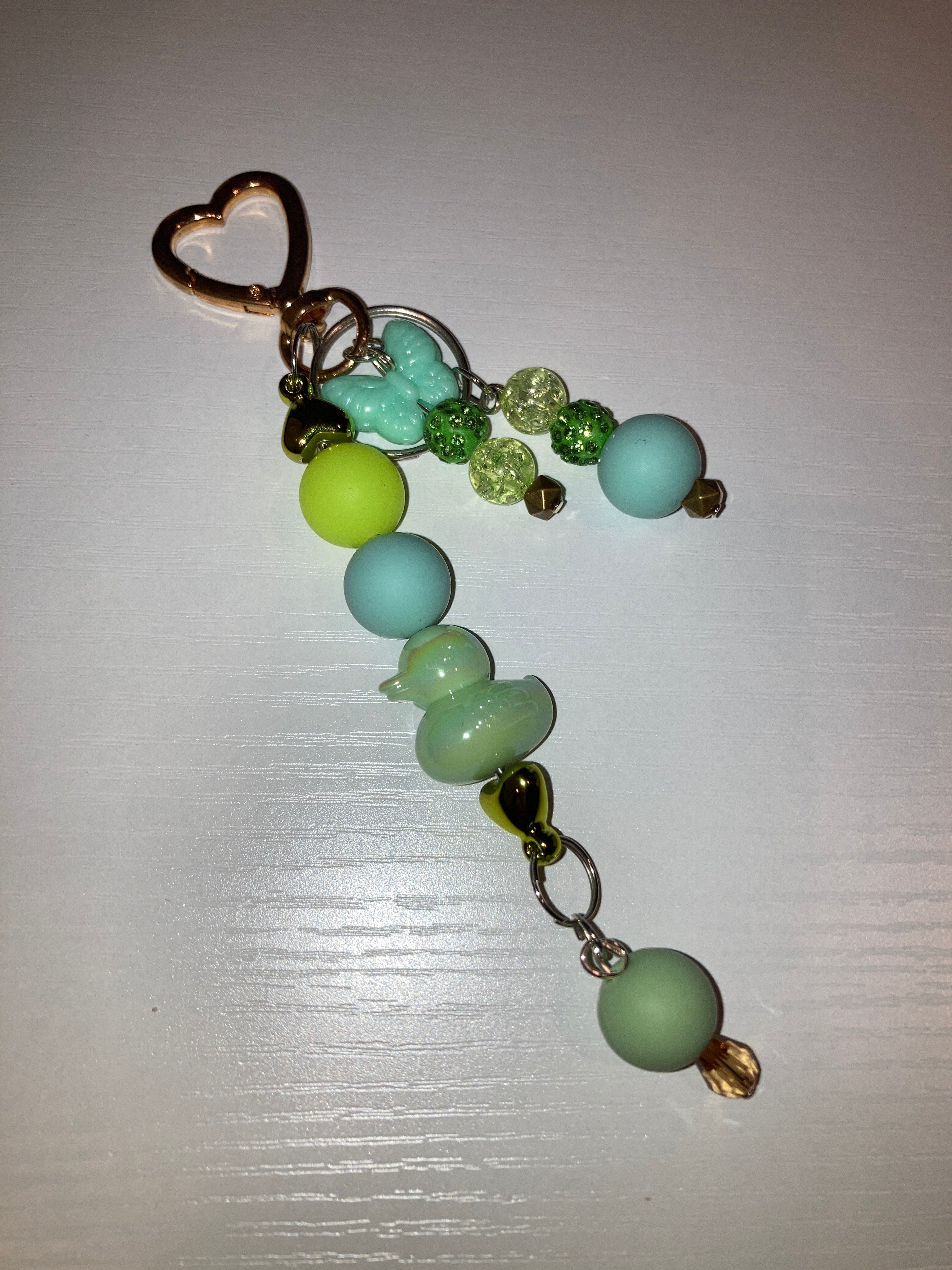 A beaded keychain or bag charm with pastel light blue, mint green, and lime green beads, clear faceted beads, a small gold heart charm, and a gold-tone metal ring, shown on a white background.