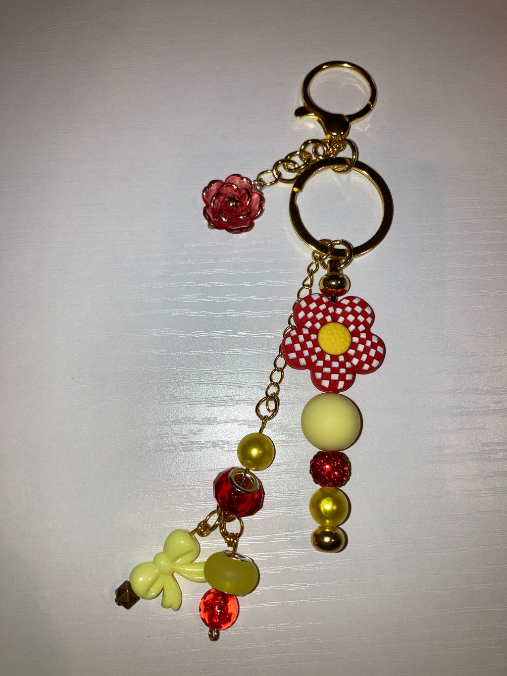 A decorative gold-tone keychain with a lobster clasp, split key ring, chain links, a red-and-white polka dot flower charm, round beads in pale yellow and red, a small red rose charm, and a pale yellow butterfly charm, displayed on a light gray background.