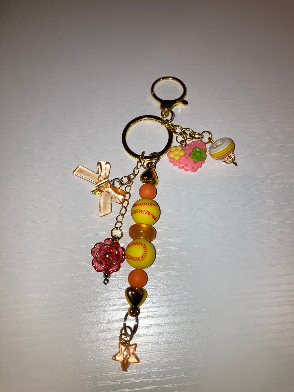 A decorative keychain featuring yellow and orange baseball-themed beads, a pink cherry charm, a gold cross, a red flower charm, and a star charm on a gold chain.