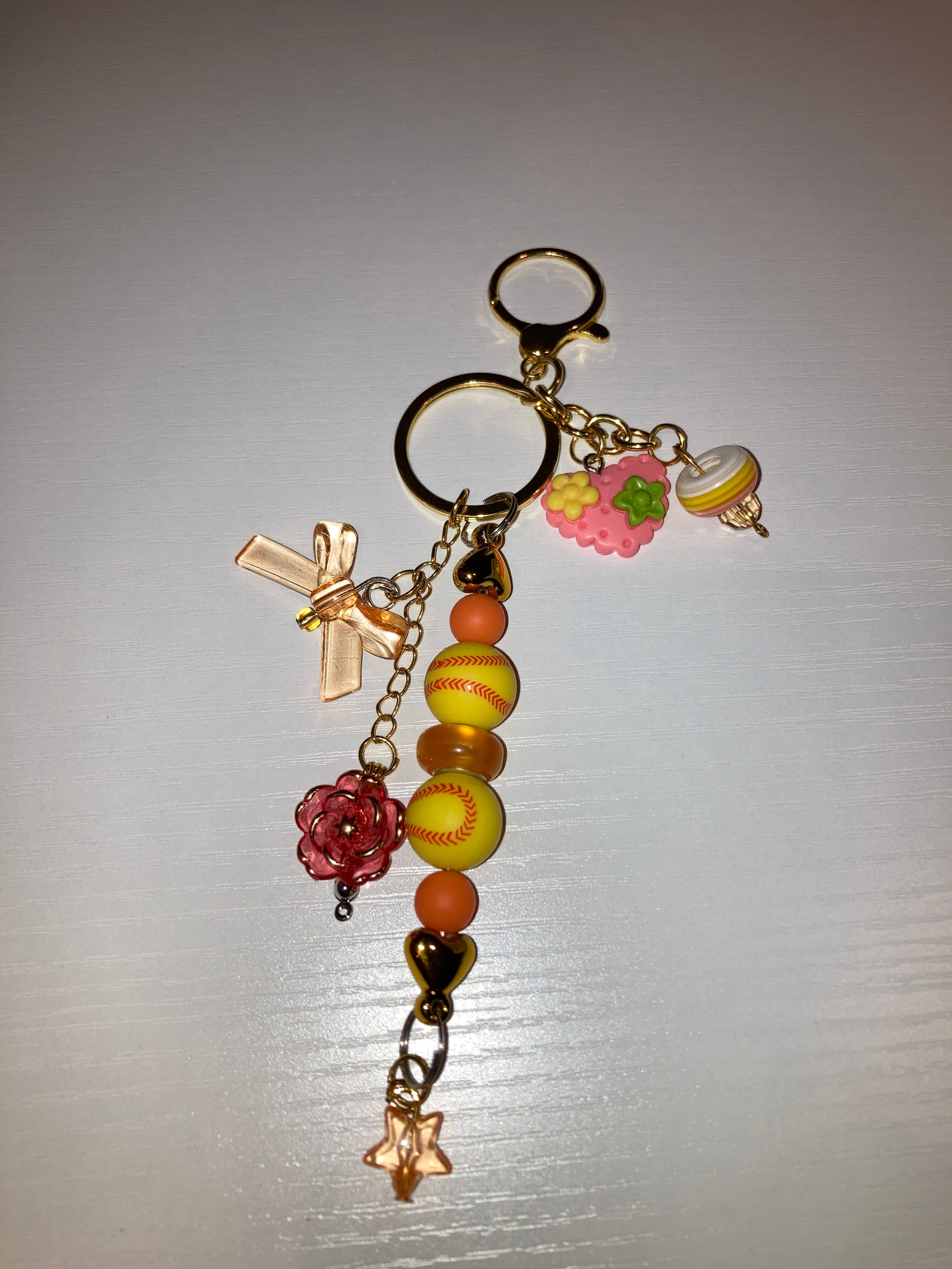 A decorative keychain featuring yellow and orange baseball-themed beads, a pink cherry charm, a gold cross, a red flower charm, and a star charm on a gold chain.
