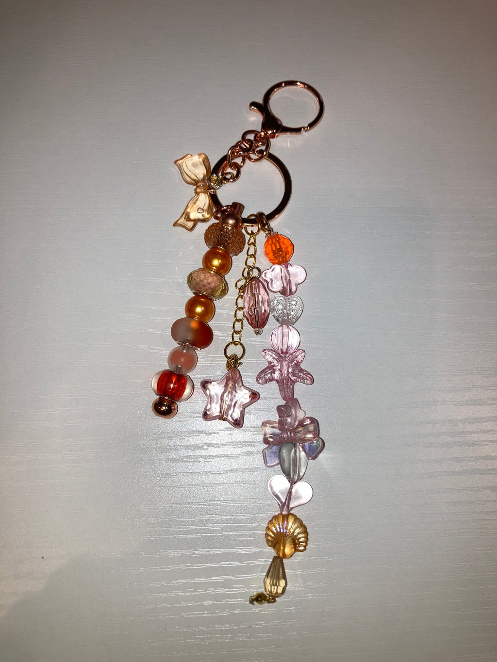 A handmade keychain featuring a rose gold key ring, a gold chain, translucent pink star-shaped beads, a gold flower charm, and a dangling beaded strand with orange, brown, and pink beads.