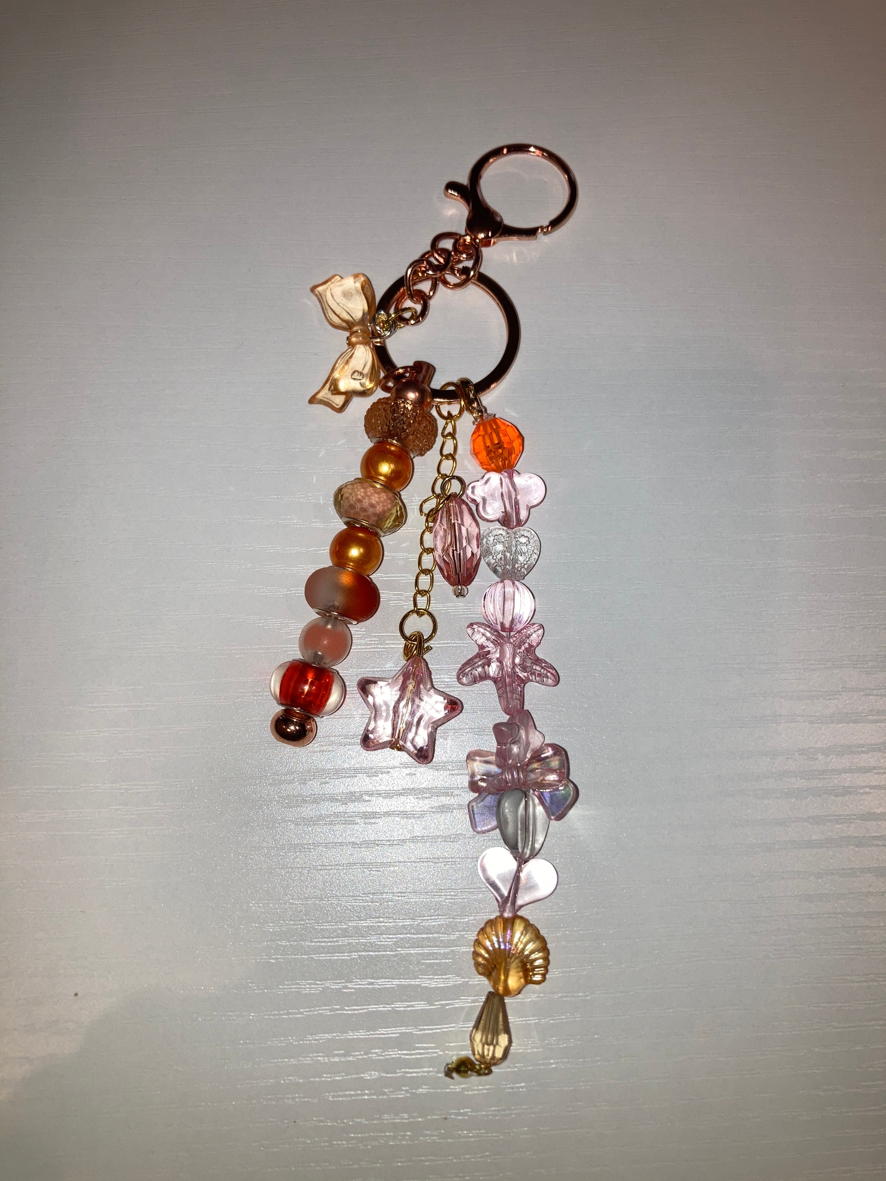 A handmade keychain featuring a rose gold key ring, a gold chain, translucent pink star-shaped beads, a gold flower charm, and a dangling beaded strand with orange, brown, and pink beads.