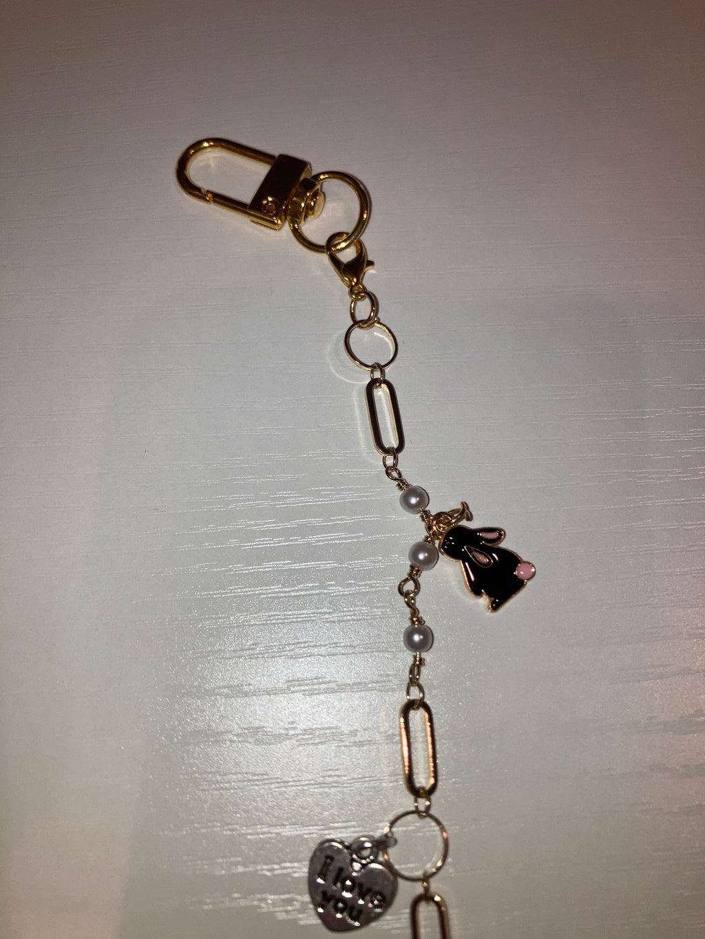 A gold-tone chain bracelet with a lobster clasp, paperclip links, small pearl-like beads, a black bunny charm, and a silver heart charm, displayed on a light-colored surface.