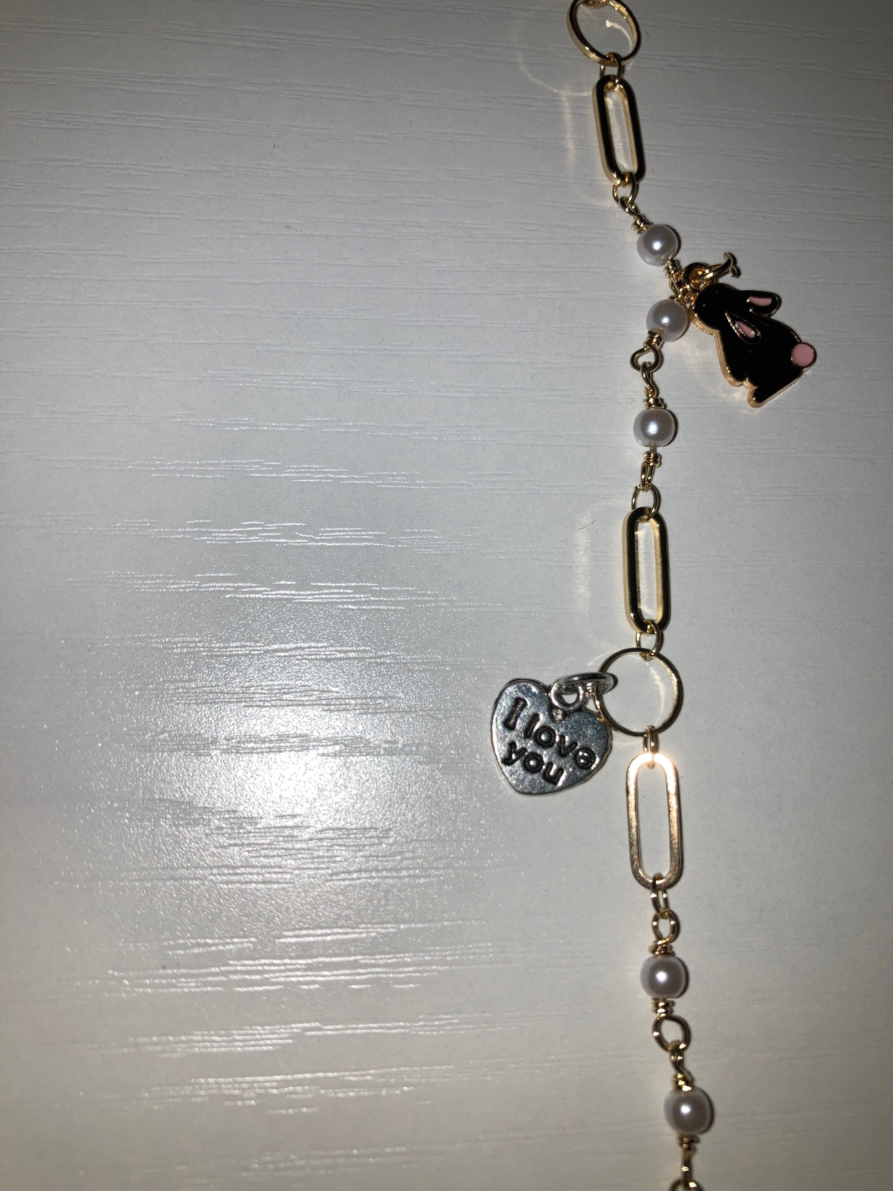 A gold-tone chain bracelet featuring faux pearl beads, elongated oval links, a small black-and-white cat charm, and a silver-tone heart-shaped charm with printed eyes and teeth, displayed on a light gray surface.