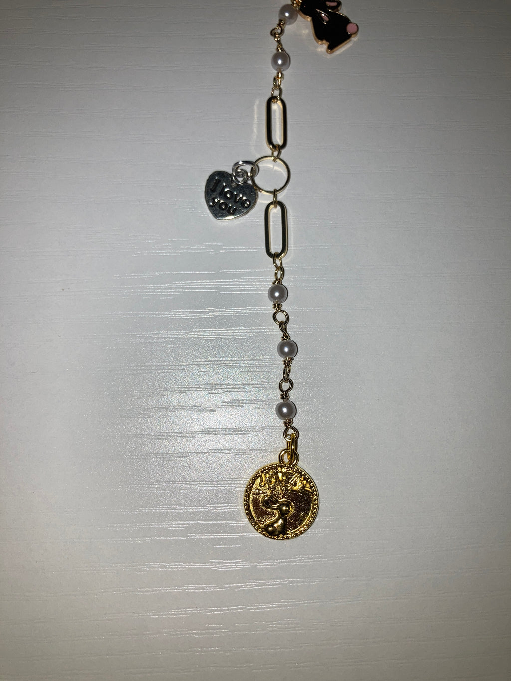 A gold-tone chain bracelet featuring faux pearl beads, elongated oval links, a small black-and-white cat charm, and a silver-tone heart-shaped charm with printed eyes and teeth, displayed on a light gray surface.
