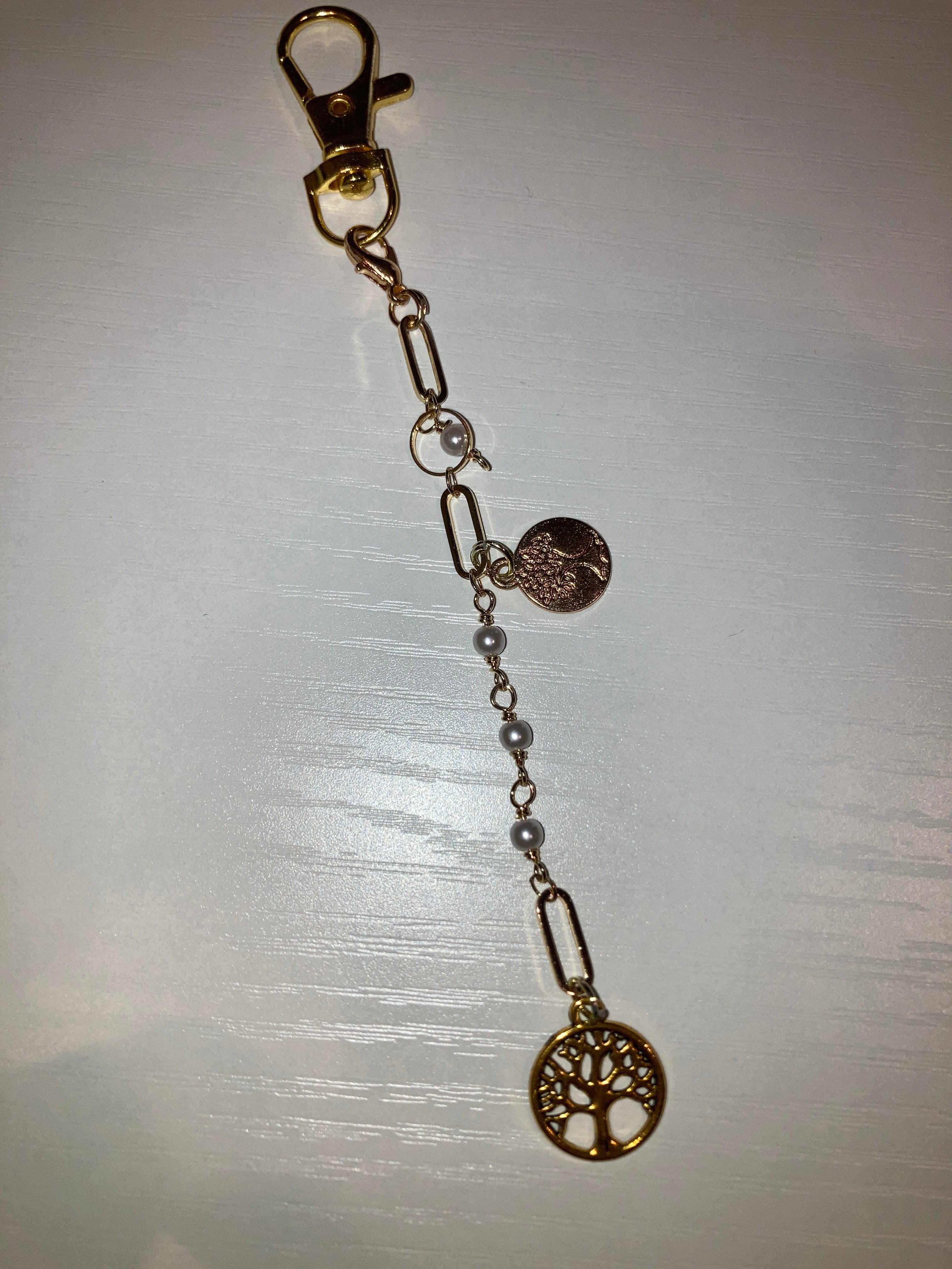 A gold-tone keychain accessory featuring a lobster clasp, chain links, small pearl-like beads, a round engraved charm, and a tree-of-life pendant, displayed on a light gray surface.