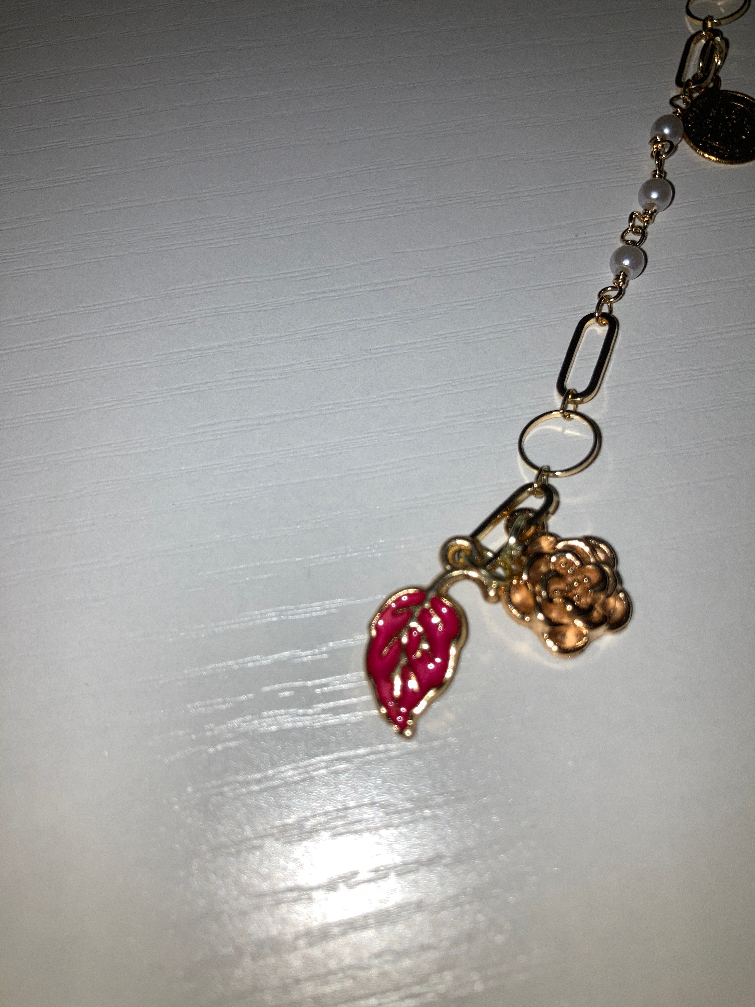 A close-up of a gold-tone chain bracelet featuring a small gold flower charm and a pink enamel lip charm, resting on a white surface.