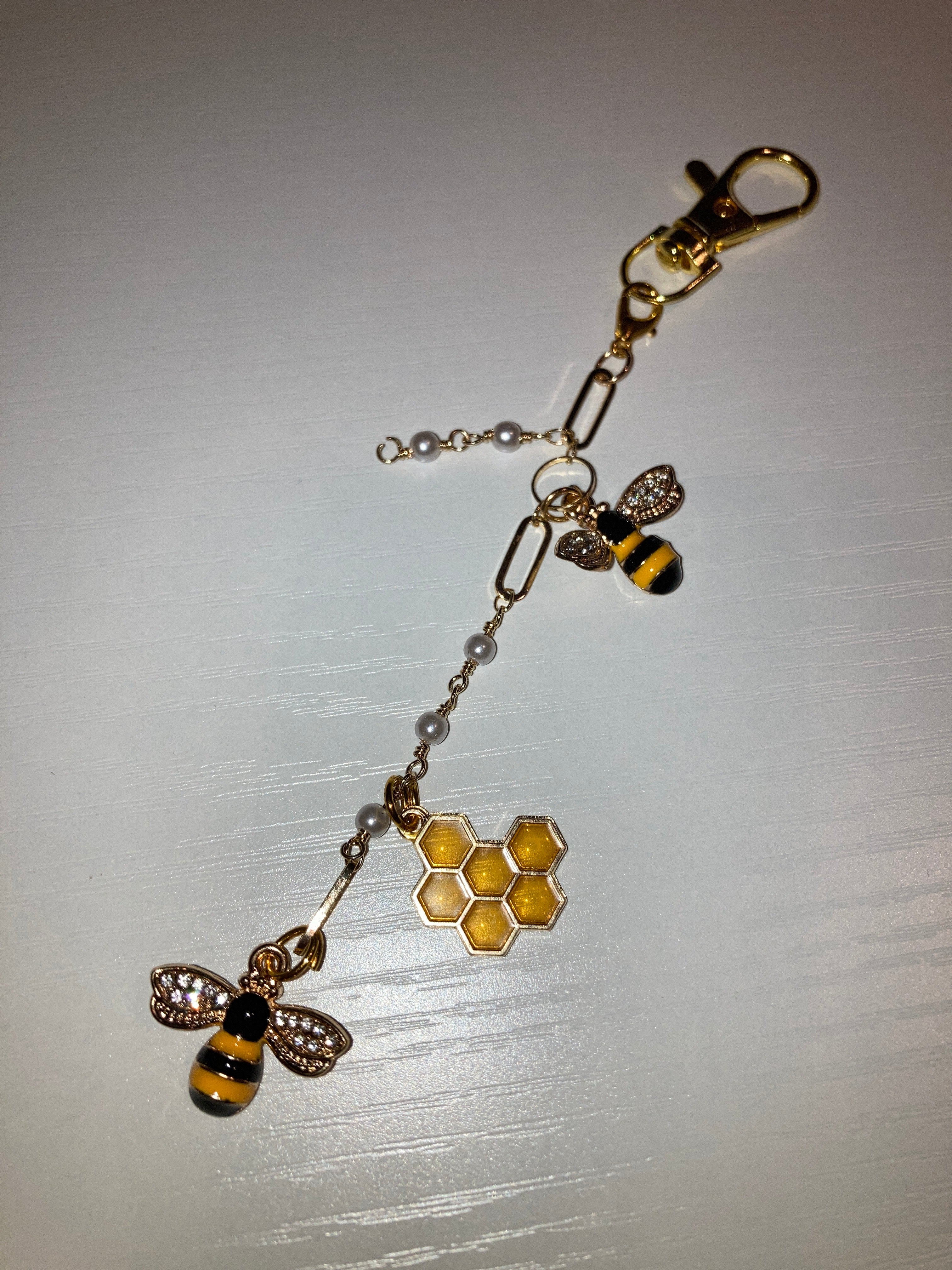 A gold-tone beehive keychain featuring two bumblebee charms, a honeycomb pattern, and small pearl beads on a white background.