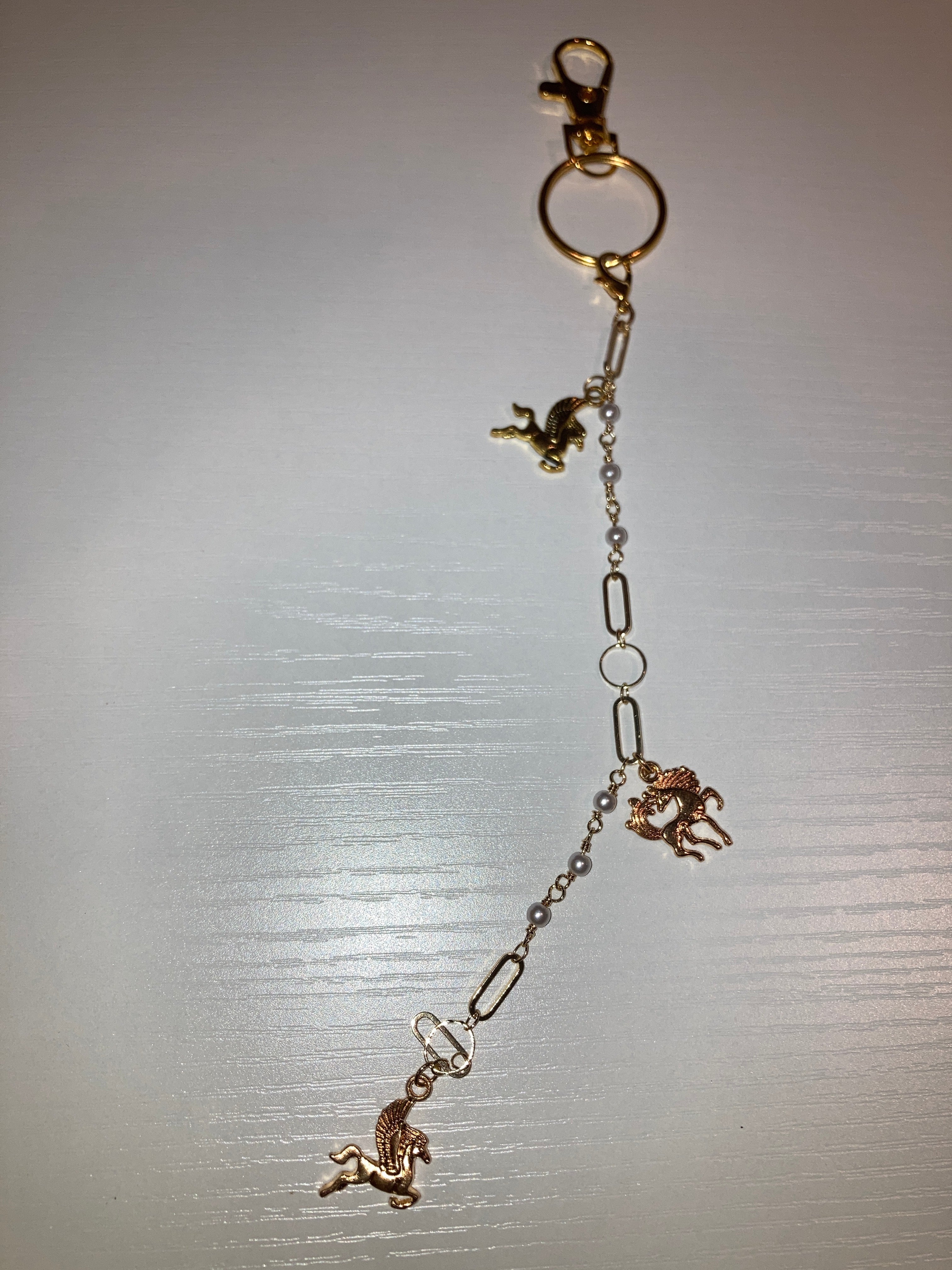 A gold-toned keychain or bag charm with a lobster clasp, split ring, and dangling chain links, decorated with four small horse charms in a mix of gold and rose gold tones, shown on a light gray background.