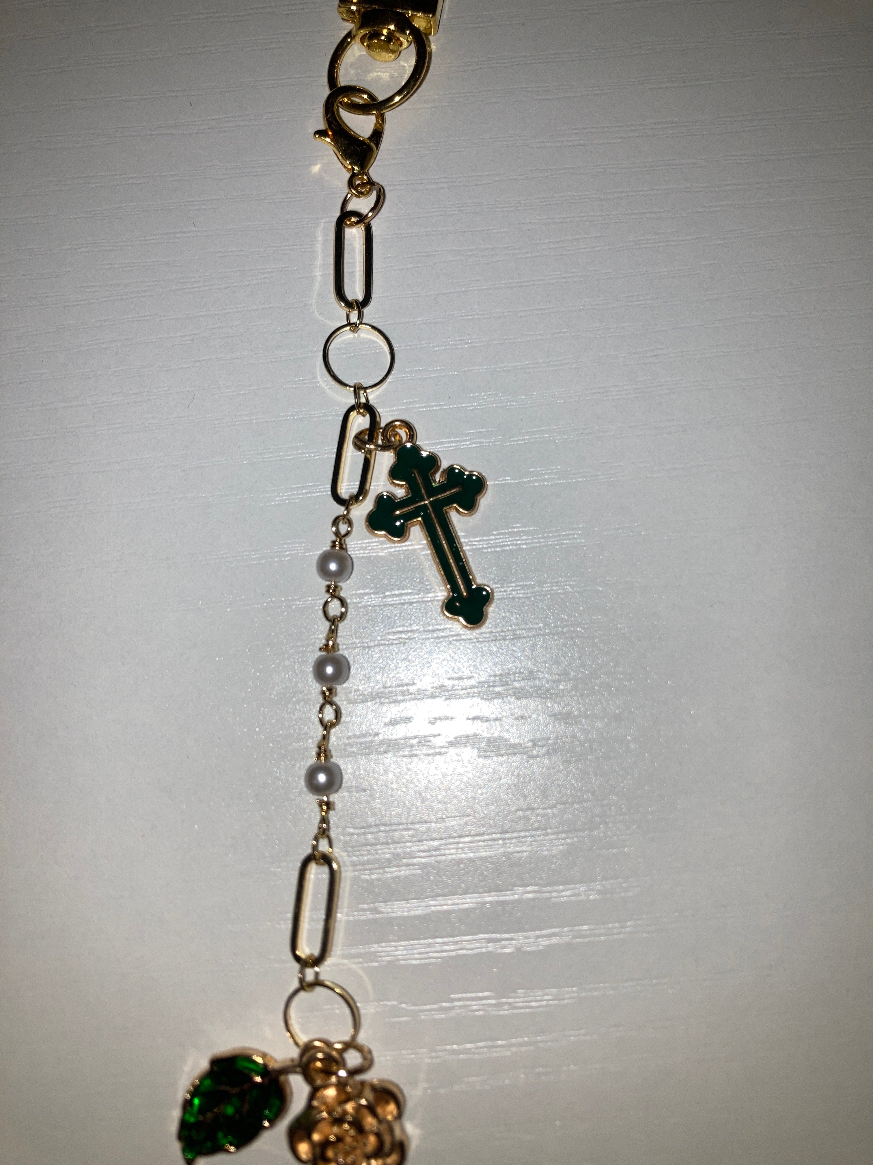 A gold-toned chain bracelet featuring a small green cross charm, white pearl beads, and a green faceted gemstone charm, displayed on a light gray background.