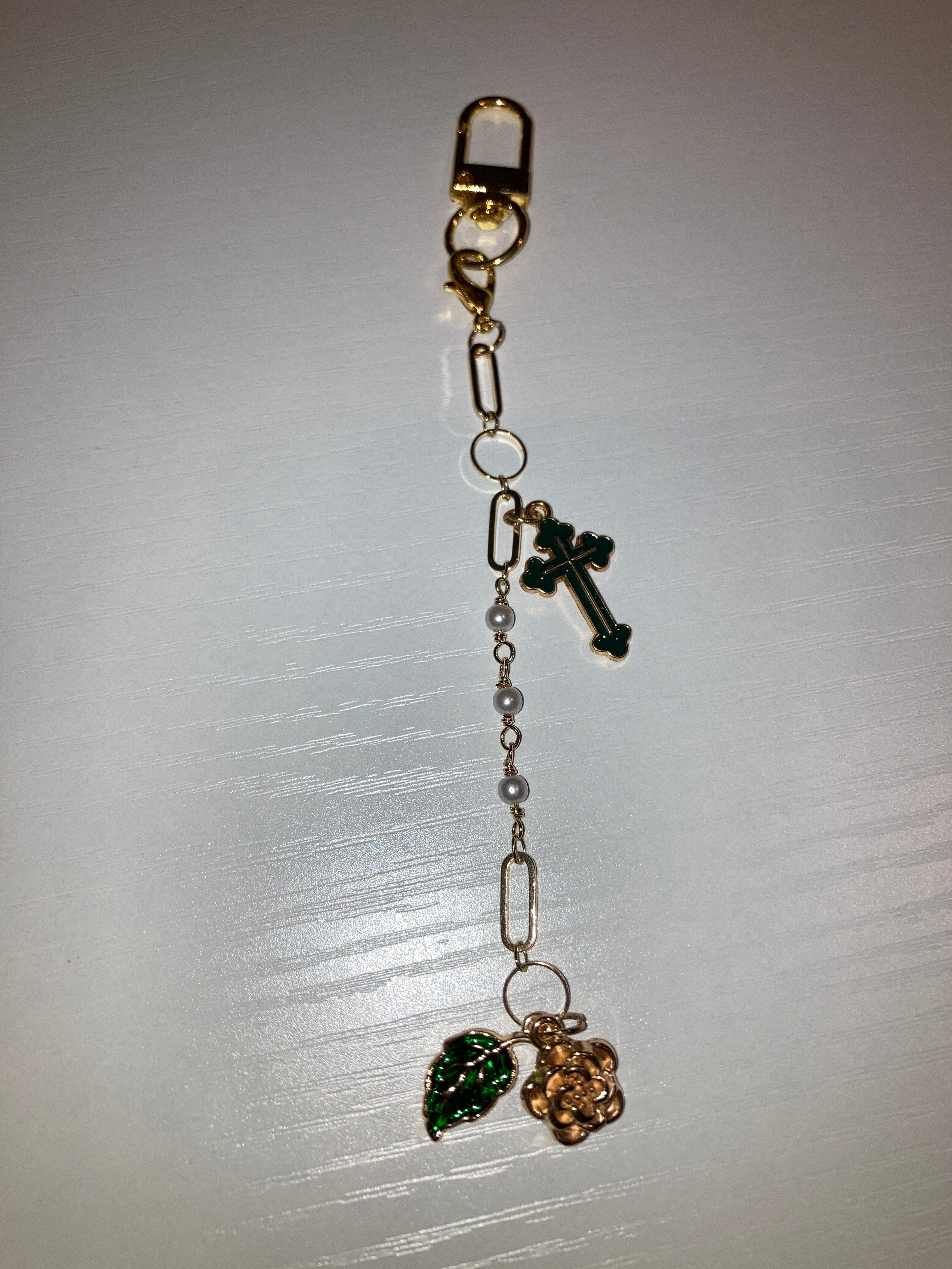 A gold-toned chain bracelet featuring a small green cross charm, white pearl beads, and a green faceted gemstone charm, displayed on a light gray background.