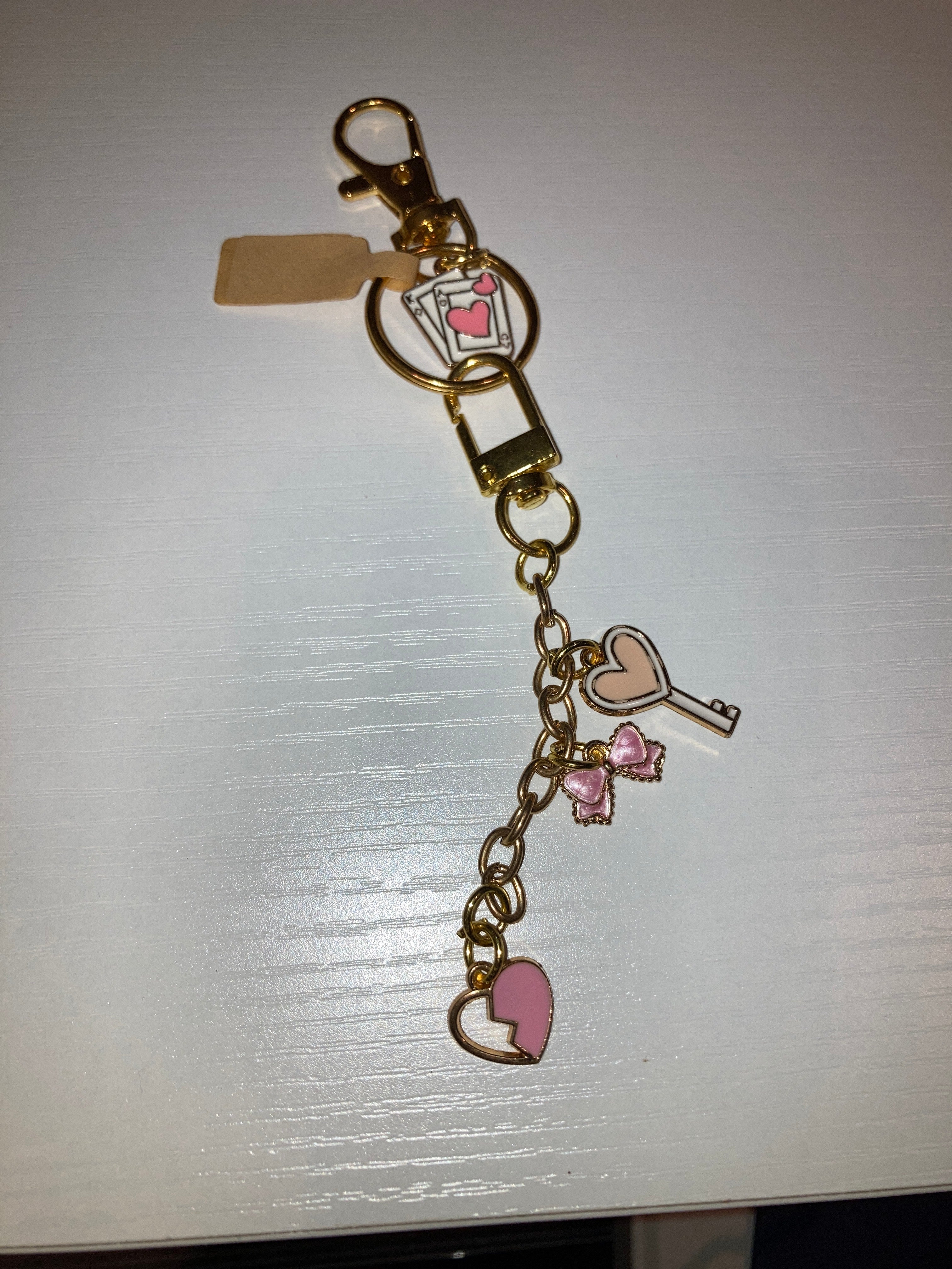 A gold-tone metal keychain accessory with a lobster clasp, a heart-shaped charm, a key-shaped charm, two small pink bow charms, and a pink heart charm, displayed on a white surface.